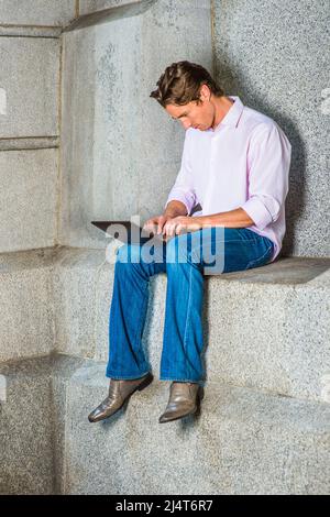 Mann, Der Draußen Arbeitet. Ein junger Geschäftsmann sitzt in einem hellrosa, langärmeligen Hemd, blauen Jeans und Lederschuhen lässig gegen eine Betonwa Stockfoto