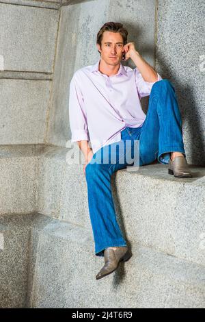 Mann, der am Telefon anruft. In einem hellrosa Langarmshirt, einer blauen Jeans und Lederschuhen sitzt ein junger Geschäftsmann lässig gegen einen Betonklotz Stockfoto