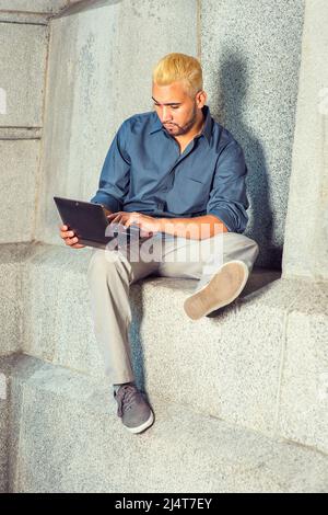 Junger Mann, Der Draußen Arbeitet. Ein blaues Hemd, graue Hosen, Freizeitschuhe, ein junger Mann mit Bart, gelbe Haare sitzen an einer Betonwand und schauen Stockfoto