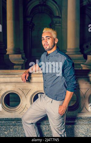 Porträt eines jungen Arbeiters. In einem schwarz gemusterten Hemd, einer gelben Hose, steht ein junger Mann mit Bart, gelben Haaren an einem Geländer auf dem Büro h Stockfoto