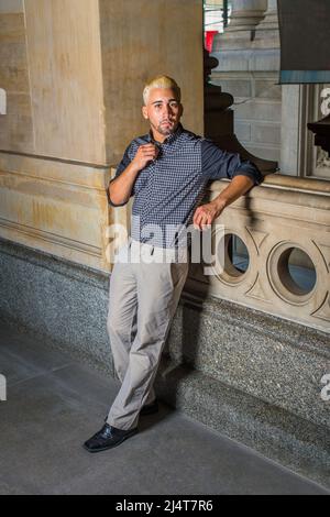 Porträt eines jungen Arbeiters. In einem schwarz gemusterten Hemd, einer grauen Hose, Lederschuhen, steht ein junger Mann mit Bart, gelben Haaren an einem Geländer Stockfoto