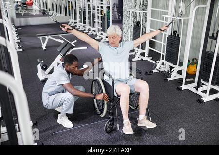 Porträt eines afroamerikanischen Rehabilitationstherapeuten, der älteren Mannes im Rollstuhl bei körperlichen Übungen im Fitnessstudio assistiert Stockfoto