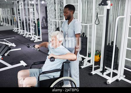 Porträt eines afroamerikanischen Rehabilitationstherapeuten, der älteren Menschen im Rollstuhl im Fitnessstudio assistiert, Kopierraum Stockfoto