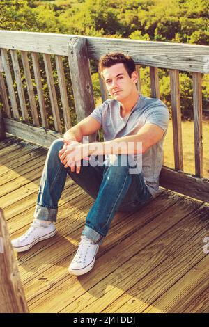 Mann, Der Sich Draußen Entspannt. In einem grauen T-Shirt, Jeans und weißen Sneakers sitzt ein junger, hübscher Kerl auf dem Holzboden, zurück gegen den Zaun in einem remo Stockfoto