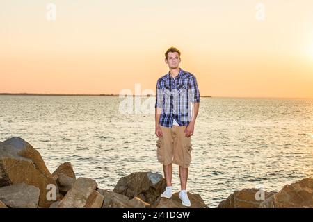 Mann wartet auf dich bei Sonnenuntergang. In einem weißen, blau gemusterten Hemd, einer gelben Hose, weißen Turnschuhen, steht ein junger, gutaussehender Kerl auf Felsen bei einer Bucht, Stockfoto