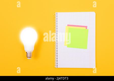 Ein Ideenkonzept - leuchtende Energiesparlampe und EIN Notizblatt mit Haftnotizen auf gelbem Tisch. Draufsicht, Flach Liegend, Leere Vorlage Stockfoto