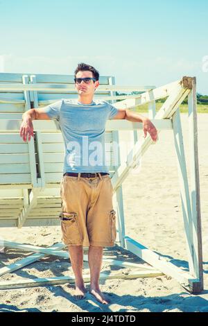 Mann Sommer Casual Fashion. In einem grauen T-Shirt, einer lässigen kurzen Hose, einer Sonnenbrille und auf einem Holzstock ruhenden Armen steht ein junger, gutaussehender Kerl zur Verfügung Stockfoto