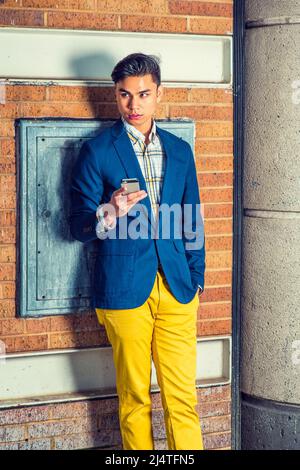 Warten Auf Ihren Anruf. Dressing in blauen Blazer, gelbe Hosen, Hand hält ein Handy. Ein junger gutaussehender Kerl steht an einer braunen Ziegelwand, Stockfoto