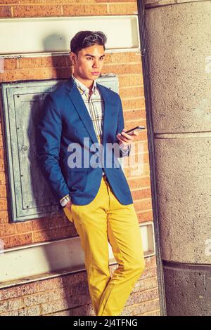 Warten Auf Ihren Anruf. Dressing in blauen Blazer, gelbe Hosen, Hand hält ein Handy. Ein junger gutaussehender Kerl steht an einer braunen Ziegelwand, Stockfoto