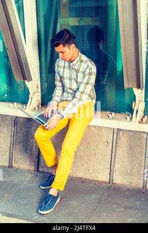 Mann, der draußen arbeitet. In einem weißen, gelb gemusterten Hemd, einer gelben Hose und blauen Sneakers sitzt ein junger, hübscher College-Student an der Metallst Stockfoto