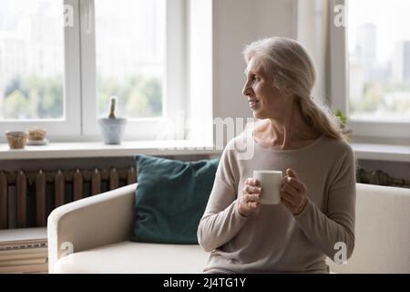 Verträumte ältere alleinerziehende Frau hält den Becher mit Blick aus dem Fenster Stockfoto