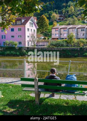 Ein Reisender mit Rucksack sitzt auf einer grünen Bank am Ufer des Doubs River in Saint Ursanne Stockfoto