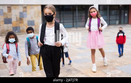 Schulmädchen in medizinischer Maske stehend auf der Straße, Kinder im Hintergrund Stockfoto