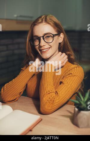 Wunderschöne rothaarige Frau, die sich zu Hause entspannt und ein Buch am Tisch liest. Moderner Lifestyle. High School University College Konzept Stockfoto