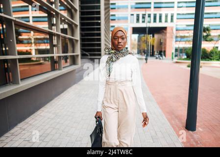 Selbstbewusste afroamerikanische Frau in stilvoller leichter Kleidung und Hijab, die an modernen Bürotürmen vorbeigeht und wegschaut Stockfoto
