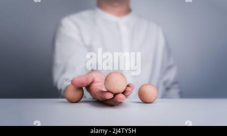 Die hölzerne Kugel in der Hand des Geschäftsmannes. Eine auserwählte Holzkugel, die von einem Geschäftsmann aus drei Kugeln auf einem weißen Tisch ausgewählt wurde. Das Konzept der besten Wahl Stockfoto
