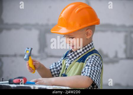 Kind in Harthut mit Hammer. Kleines Kind hilft mit Spielzeugwerkzeugen auf der Baustelle. Kinder mit Bauwerkzeugen. Bauarbeiter. Reparieren Stockfoto