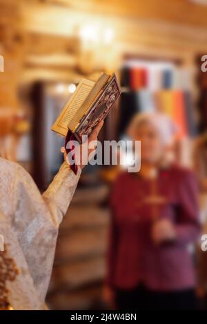 Beschnittenes Foto des Priesters, der ein dekoriertes Gebetsbuch hält, während der Zeremonie vor der Frau, die das Kreuz hält, betet. Stockfoto