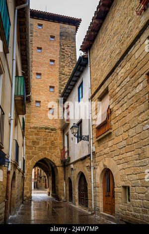 Mittelalterliche Steinhäuser und Torbogen an einer Straße in Olite, Spanien, die an einem regnerischen Tag für eine prächtige Burg des Königlichen Palastes mit Pfützen auf Kopfsteinpflaster berühmt ist Stockfoto
