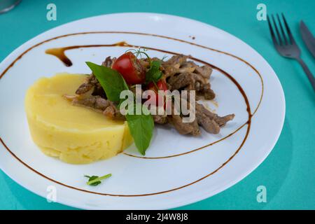 Fleischbällchen mit Kartoffelpüree auf einem weißen Teller. Fleisch in saurer Rahmsoße. Tomaten und Basilikum. Essen im Restaurant. Mittagspause. Medaillons. Stockfoto