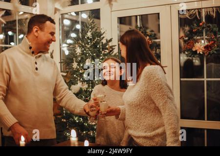 Familie trinkt hausgemachten Eiertang am Weihnachtsbaum Spaß haben. Schöne Frau und Mann mit tauhter zusammen. Weihnachtsbaum auf dem Hintergrund. Hygge c Stockfoto