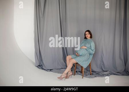 Attraktive Schwangere in grünem Kleid sitzt auf Holzstuhl auf blauem Hintergrund. Mutterschaft und Gesundheitskonzept Stockfoto