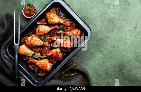 Gebackene Hähnchenbeine, Drumsticks mit süßen Feigen und Zwiebeln in balsamischer Marinade auf schwarzem Backblech, rostiger grüner Tischhintergrund, Draufsicht Stockfoto