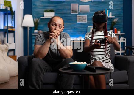 Kaukasischer Mann beobachten afroamerikanische Frau spielen VR-Spiel. Ehemann sitzt neben Frau mit Virtual-Reality-Brille. Ein vielfältiges Paar genießt Wearable-Technologie und Simulationsspiele Stockfoto