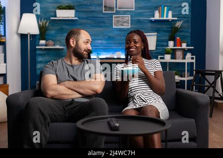 Glückliches interrassisches Paar, das ein angenehmes Gespräch hat, während es zu Hause auf dem Sofa sitzt. Entspannter Mann und Frau verbinden sich und haben eine gute Zeit im Wohnzimmer, während Sie Snacks teilen Stockfoto