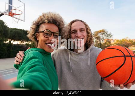 Selfie einer lateinerin und eines kaukasischen Mannes, die glücklich lächeln, nachdem sie in einem Basketballplatz im Stadtpark gespielt haben, Konzept der Freundschaft und des städtischen Sports in der St. Stockfoto