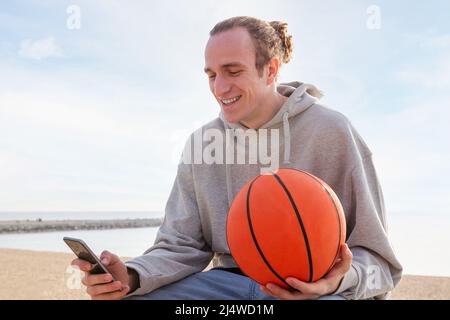 Junger kaukasischer Mann mit Kapuzenpullover und einem lachenden Basketball mit seinem Mobiltelefon, Lifestyle- und Kommunikationstechnologie, Copy Space fo Stockfoto