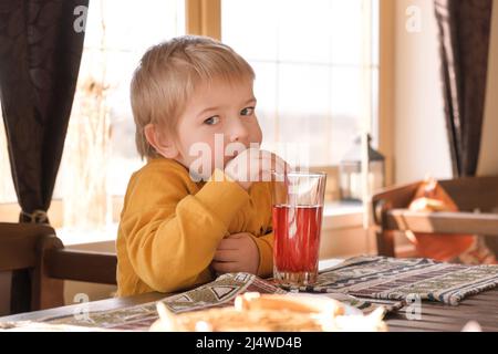 Kind Junge mit blonden Haaren Baby trinkt Limonade im Café. 3 Jahre altes Kind trinkt roten Saft aus Gläsern durch Trinkhalme. Junge Kleinkind in einem Restaurant. Stockfoto