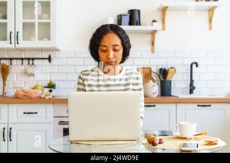 Fokussierte afroamerikanische Frau, die aus der Küche auf einem Laptop arbeitet. Konzept eines Remote-Büros zu Hause Stockfoto