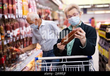 Reife Frau in Maske und Handschuhen mit Covid Schutz nimmt Salami in Fleisch Abschnitt des Supermarkts Stockfoto