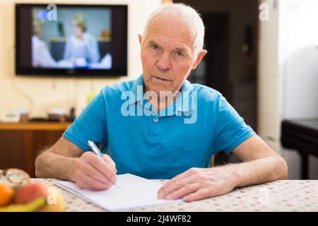 Fokussierter älterer Mann, der zu Hause sitzt und Papiere liest und unterschreibt Stockfoto