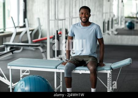 Porträt eines afroamerikanischen Mannes, der in der Rehabilitationsklinik auf einem Massagebett sitzt und die Kamera anlächelt Stockfoto