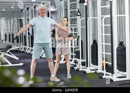 Ganzkörperportrait eines älteren Mannes, der in der Rehabilitationsklinik körperliche Übungen macht, wobei eine weibliche Therapeutin assistiert Stockfoto