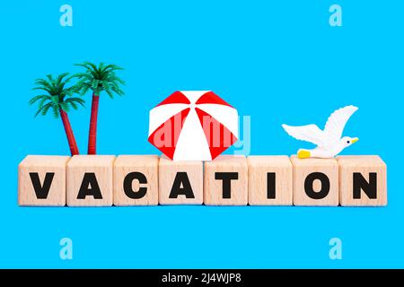 WORTURLAUB aus Buchstabenblöcken, Spielzeugpalme, Sonnenschirm und einer Möwe auf blauem Hintergrund isoliert. Kreatives Sommerurlaubs-Konzept. Stockfoto