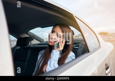 Geschäftsfrau, die während der Fahrt mit dem Taxi telefoniert. Frau, die mit Laptop und Telefon auf dem Rücksitz des Autos sitzt Stockfoto