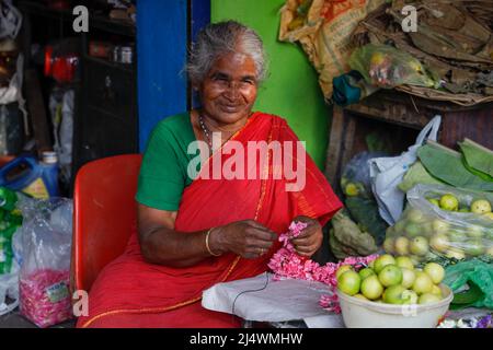 Ältere Frau, die am Straßenrand sitzt und Blumengirlanden macht und Obst in Trichy, Tamil Nadu, Indien, verkauft Stockfoto