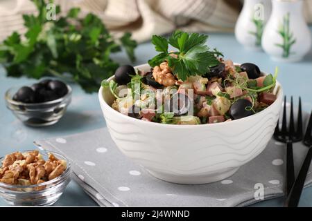 Salat mit Schinken, Ananas, schwarzen Oliven und Walnüssen in einer weißen Schüssel auf hellblauem Hintergrund Stockfoto