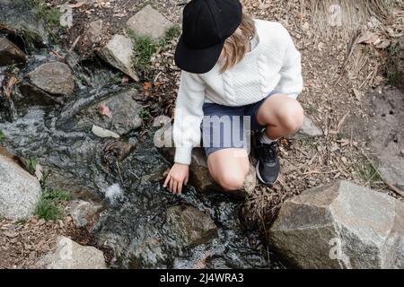 Junges Mädchen, das an einem Bach sitzt, der das Wasser berührt Stockfoto