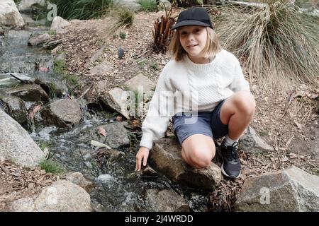 Junges Mädchen, das an einem Bach sitzt, der das Wasser berührt Stockfoto