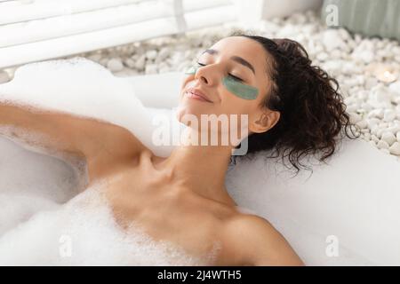Charmante junge Frau, die im Schaumbad mit Flecken unter den Augen liegt und sich zu Hause um die empfindliche Haut kümmert Stockfoto