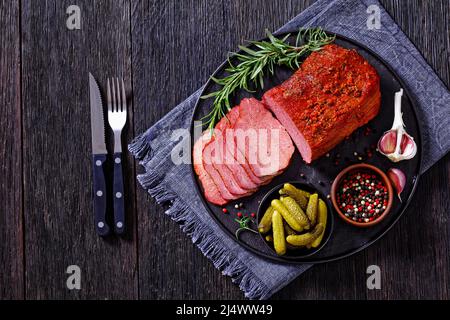 Frisch geschnittenes Rinderpastrami auf schwarzem Teller mit frischem Rosmarin, Thymen und Gurken auf dunklem Tisch, horizontale Ansicht von oben, flach liegend Stockfoto