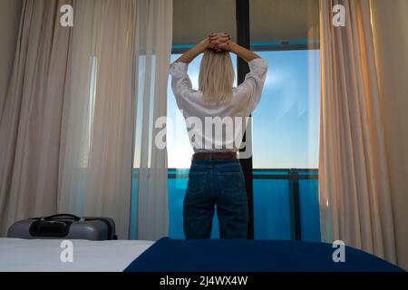 Eine junge Frau in einem weißen Hemd und einer blauen Jeans steht mit den Händen auf dem Kopf in einem Hotelzimmer am Fenster. Reisecncept und Sommerferien. Stockfoto