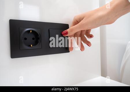 Die Nahaufnahme der Hand einer Frau mit rotem Nagellack schaltet das Licht in der Nähe des Bettes mit einem schwarzen Schalter an einer weißen Wand neben der Steckdose ein Stockfoto