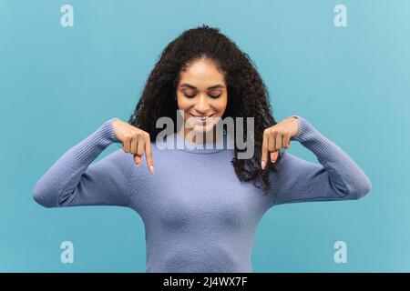Charmante junge afroamerikanische Frau zeigt die Zeigefinger nach unten und fördert Neuheit isoliert auf blauem Hintergrund, Werbekonzept Stockfoto