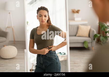 Afrikanische Dame Berührt Den Bauch Nach Gewichtszunahme In Der Nähe Des Spiegels Im Innenbereich Stockfoto