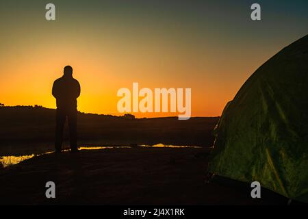 Hinterleuchteter Mann, der mit Sonnenaufgang über dem Bergschatten und einem ruhigen See mit Spiegelung am Morgen steht Stockfoto